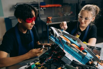 Two students work together on a robotics project in a workshop, wearing safety gear.